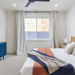 Queen bed with white bedding and layered throws, wooden headboard, and two colorful mountain art prints above it; a blue dresser and a wall-mounted TV on the opposite wall; window with gray curtains.