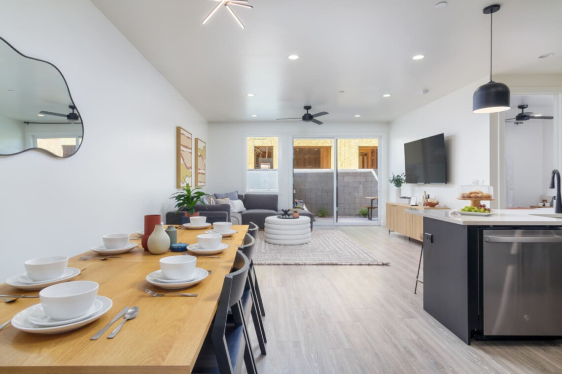 Open-concept living area with a long wooden dining table set for eight, a gray sectional, and a kitchen island with a sink and a wall-mounted TV.