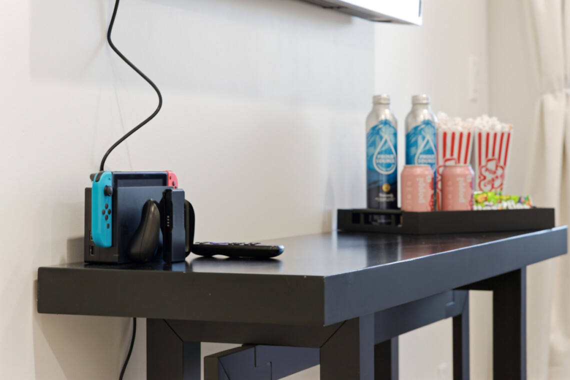 Nintendo Switch docked on a dark table with blue and red controllers, next to a remote; snack tray with water bottles and popcorn cups in the background.
