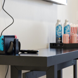 Nintendo Switch docked on a dark table with blue and red controllers, next to a remote; snack tray with water bottles and popcorn cups in the background.