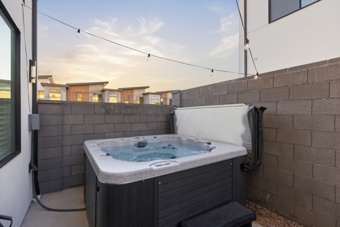 Outdoor hot tub in a concrete patio bordered by a tall gray block wall, string lights overhead at sunset/light dusk.