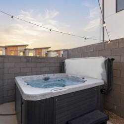 Outdoor hot tub in a concrete patio bordered by a tall gray block wall, string lights overhead at sunset/light dusk.