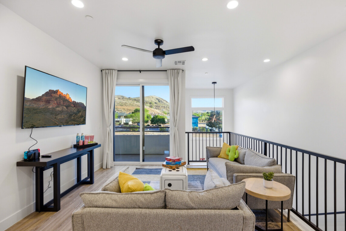 Living room with a wall-mounted TV, dark console, and beige sofas facing a sliding glass door to a balcony with hills outside.
