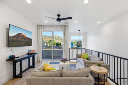 Living room with a wall-mounted TV, dark console, and beige sofas facing a sliding glass door to a balcony with hills outside.