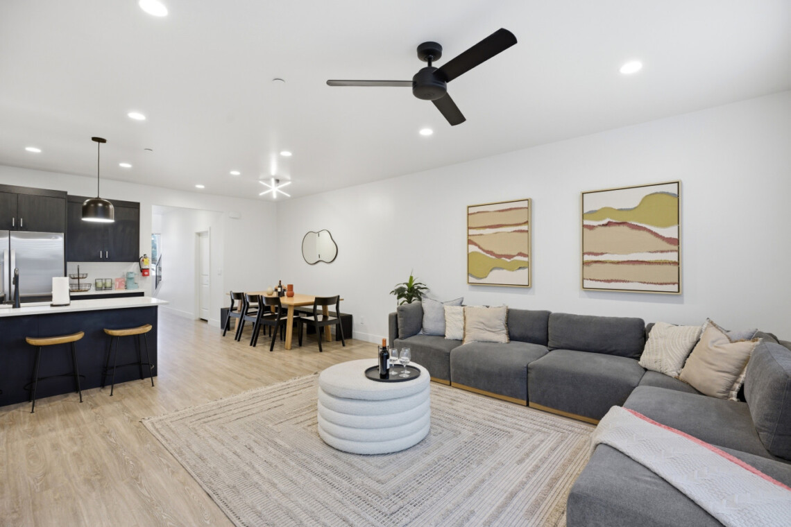 Open-plan living room and kitchen with a gray sectional sofa, abstract wall art, and a dining table in the background
