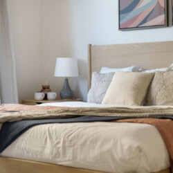 Neatly made bed with beige and gray cushions, light wood headboard, and layered blankets; bedside table with lamp and two mugs, window with white curtains nearby, and abstract art above the bed