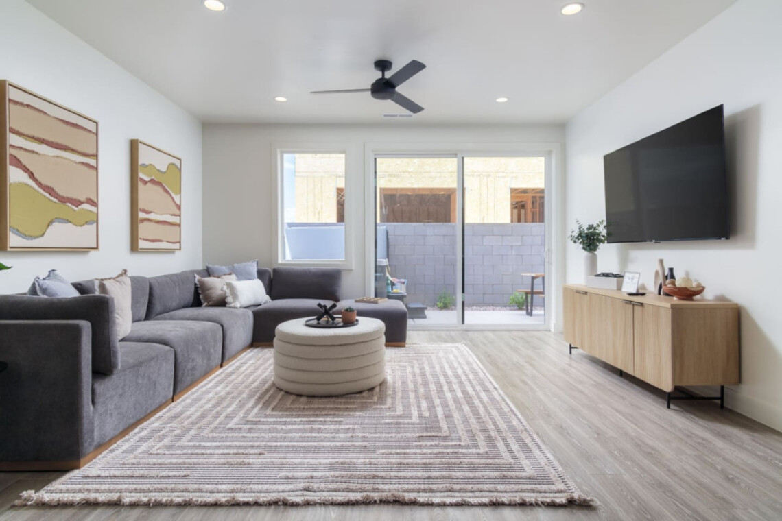 Modern living room with a gray sectional, round tufted ottoman, beige rug, wall-mounted TV, and a wooden media console.