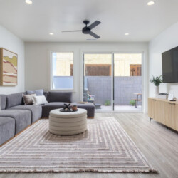 Modern living room with a gray sectional, round tufted ottoman, beige rug, wall-mounted TV, and a wooden media console.