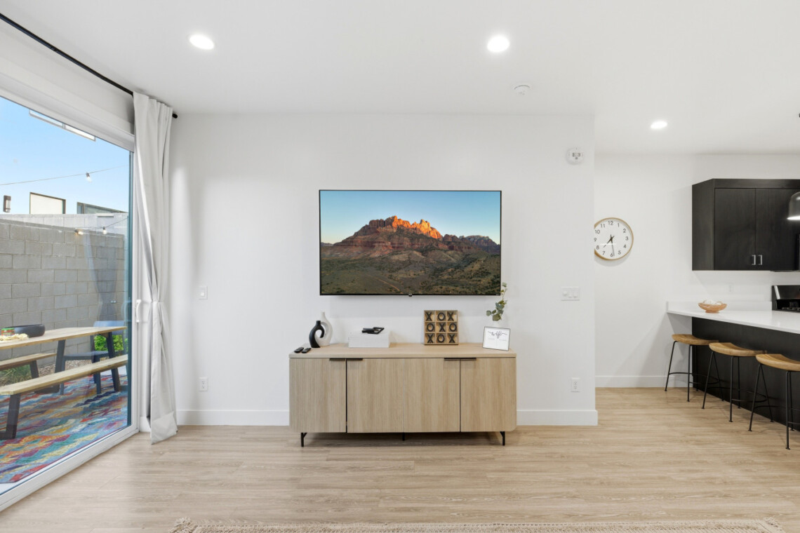 Flat-screen TV mounted above a light wood console in a bright, open living area with a sliding glass door to an outdoor patio on the left and a kitchen bar with stools on the right.