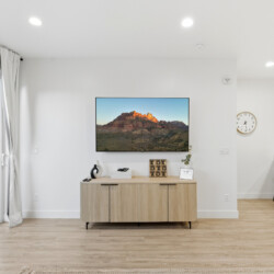 Flat-screen TV mounted above a light wood console in a bright, open living area with a sliding glass door to an outdoor patio on the left and a kitchen bar with stools on the right.