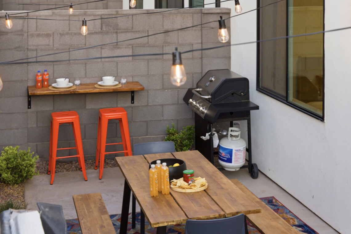 Outdoor patio with a gas grill and propane tank, a wooden table set with chips and juice bottles, and two orange stools under string lights.