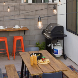 Outdoor patio with a gas grill and propane tank, a wooden table set with chips and juice bottles, and two orange stools under string lights.