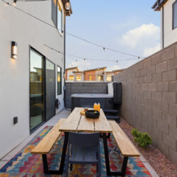 Narrow backyard patio with a wooden picnic table, benches, and string lights, next to a gray brick wall and sliding glass door.