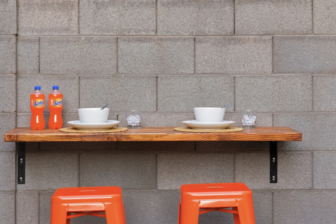 Outdoor dining setup for two on a wooden table against a gray cinder block wall, with two bowls and plates, spoons, two glasses of ice, two orange soda bottles, and two orange stools beneath.