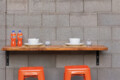 Outdoor dining setup for two on a wooden table against a gray cinder block wall, with two bowls and plates, spoons, two glasses of ice, two orange soda bottles, and two orange stools beneath.