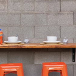 Outdoor dining setup for two on a wooden table against a gray cinder block wall, with two bowls and plates, spoons, two glasses of ice, two orange soda bottles, and two orange stools beneath.