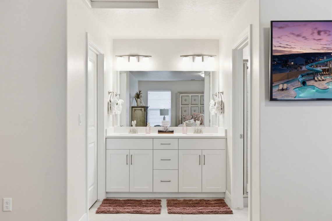 Bright, modern bathroom with a double sink vanity, large mirror, and matching white cabinets; two red bath mats on the floor and towels on the sides of the sinks.