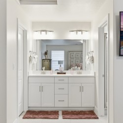 Bright, modern bathroom with a double sink vanity, large mirror, and matching white cabinets; two red bath mats on the floor and towels on the sides of the sinks.