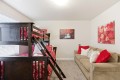 Dark wooden bunk bed with red floral bedding dominates a bright bedroom, adjoining a beige sofa with red and white cushions.