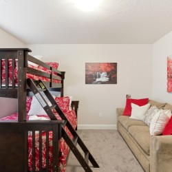 Dark wooden bunk bed with red floral bedding dominates a bright bedroom, adjoining a beige sofa with red and white cushions.