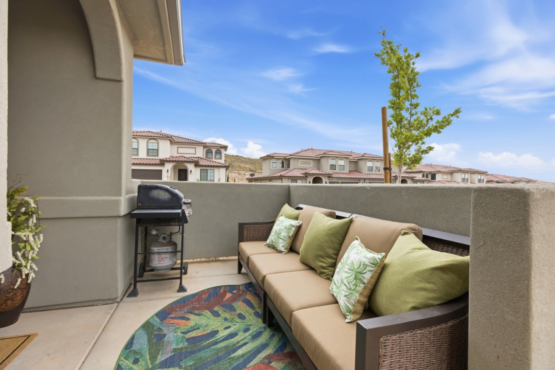 Rooftop patio with a beige sectional sofa, green pillows, a grill, and a colorful rug, overlooking townhomes.