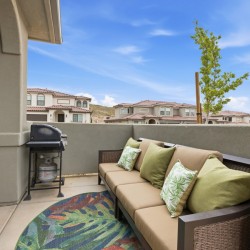 Rooftop patio with a beige sectional sofa, green pillows, a grill, and a colorful rug, overlooking townhomes.