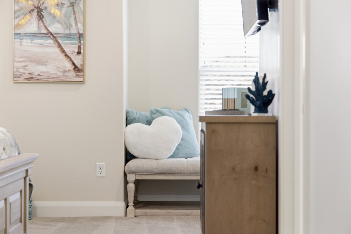 Cozy seating nook with a light beige loveseat, teal cushions, and a white heart pillow beside a wooden dresser by a window with blinds.