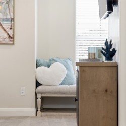 Cozy seating nook with a light beige loveseat, teal cushions, and a white heart pillow beside a wooden dresser by a window with blinds.