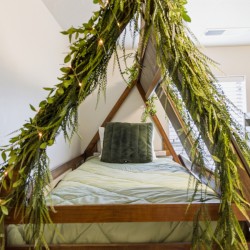 Wooden triangular bed frame under a canopy of hanging greenery with fairy lights and green bedding, creating a nature-inspired cozy bedroom atmosphere.