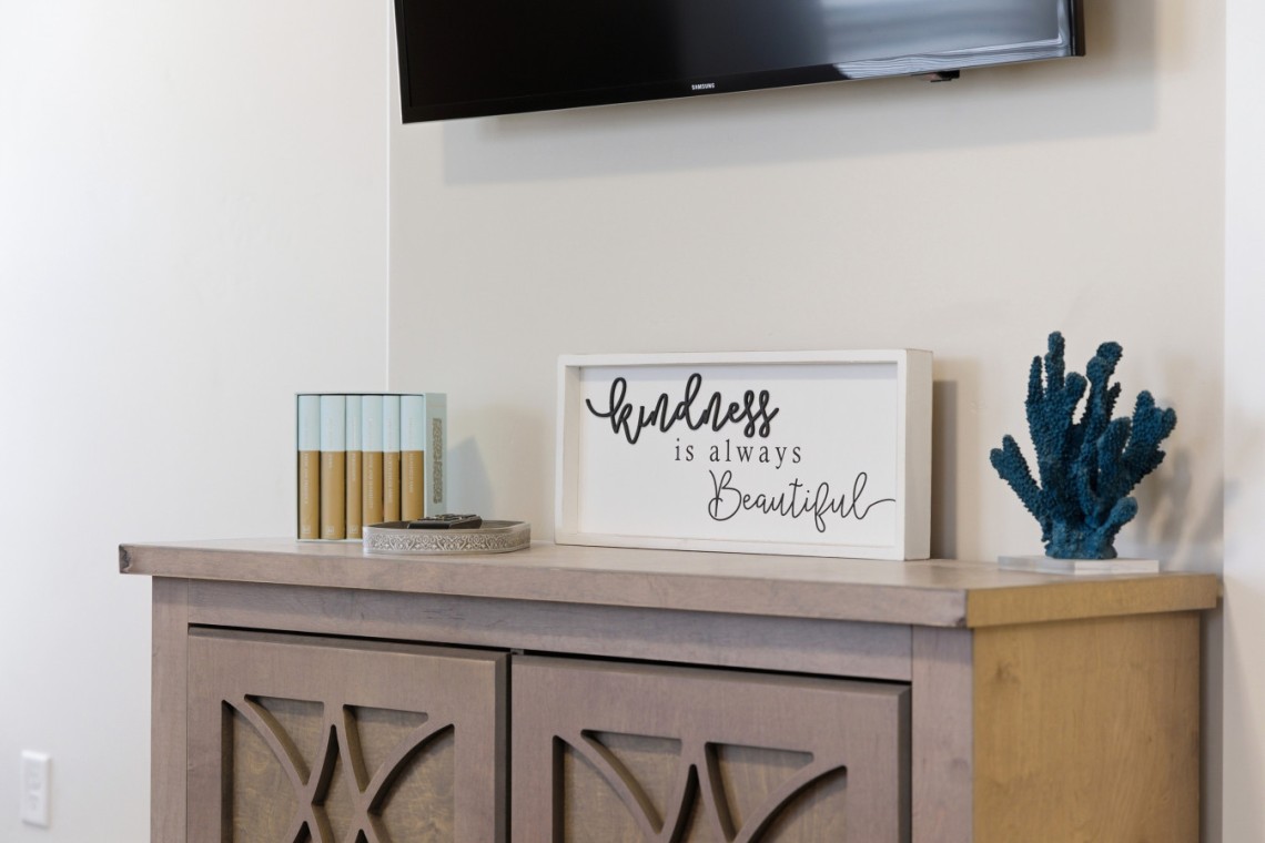 Decorative console with a framed quote reading 'kindness is always beautiful', a blue coral sculpture, and small boxed items on a light wood cabinet.