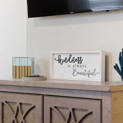 Decorative console with a framed quote reading 'kindness is always beautiful', a blue coral sculpture, and small boxed items on a light wood cabinet.