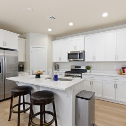 Bright white kitchen with a center island, stainless steel refrigerator, microwave, and bar stools at the island.