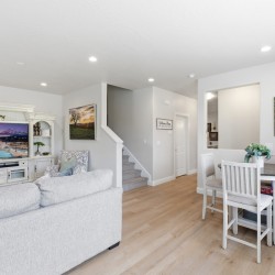 Bright open living room with a light gray sofa, white entertainment center, and a round dining table with chairs near a staircase.