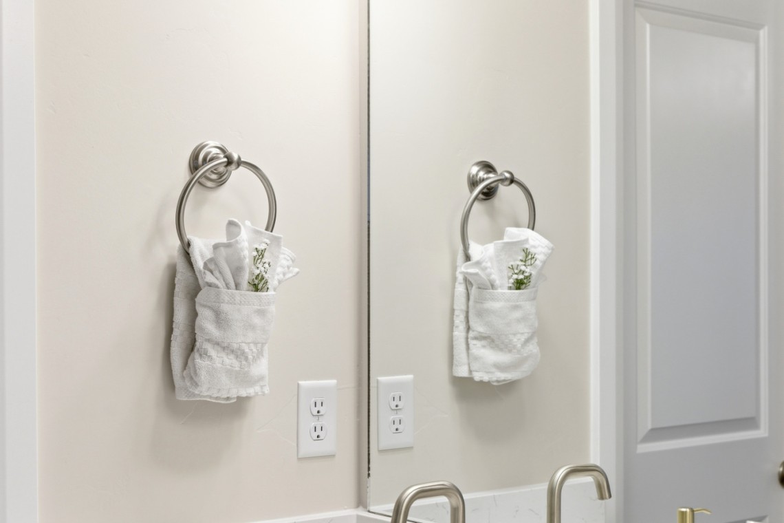 Two white towels with greenery hang from circular metal towel rings on a bathroom wall, mirrored by a split reflection of the sink area.
