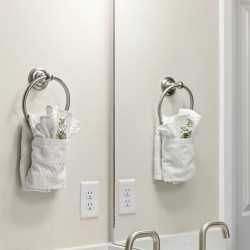 Two white towels with greenery hang from circular metal towel rings on a bathroom wall, mirrored by a split reflection of the sink area.