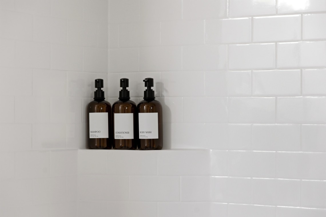 Three brown pump bottles—shampoo, conditioner, and body wash—on a tiled shower shelf against white subway tiles