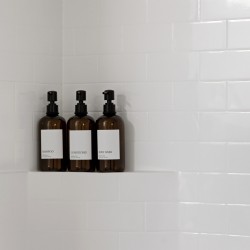 Three brown pump bottles—shampoo, conditioner, and body wash—on a tiled shower shelf against white subway tiles