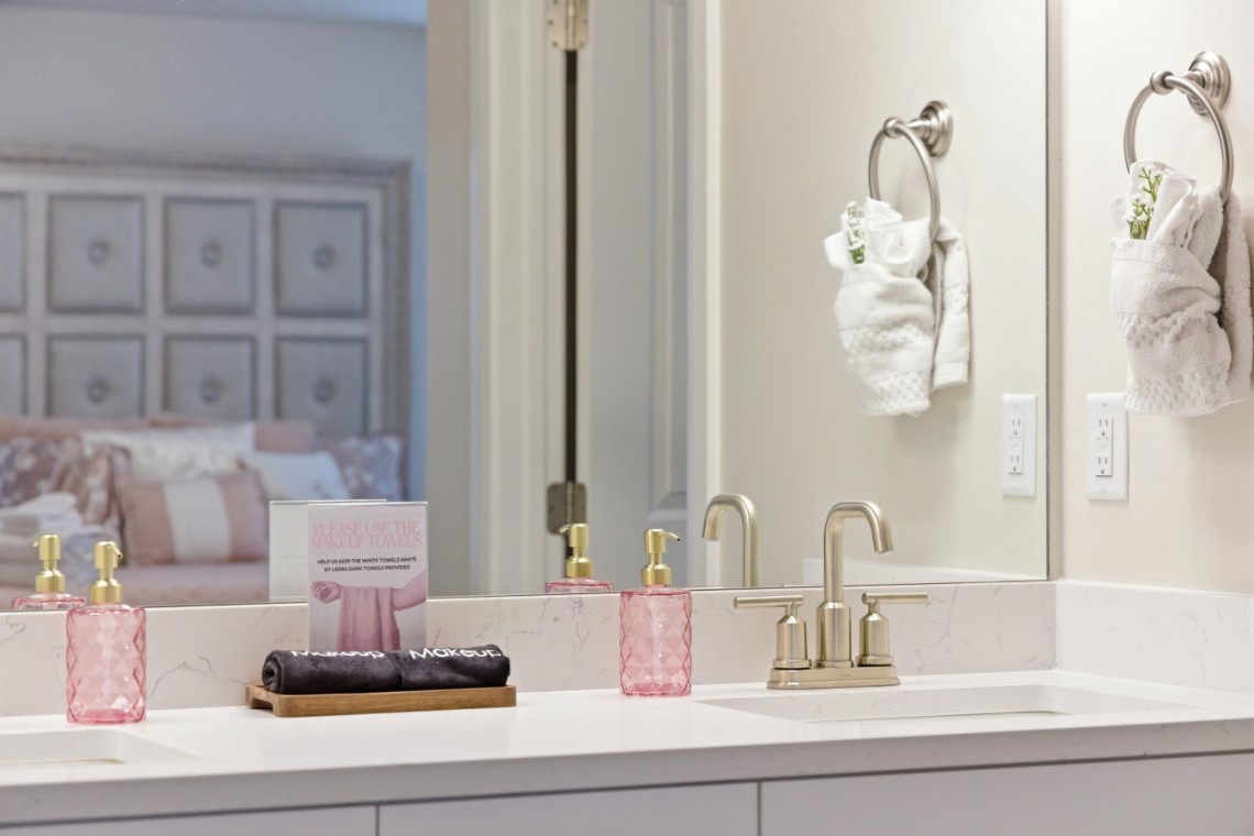 White bathroom vanity with a marble countertop, two gold faucets, and pink textured soap dispensers beside a black towel on a wooden tray; towel rings hold white towels, and a large mirror reflects a softly-lit bedroom.