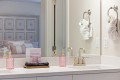 White bathroom vanity with a marble countertop, two gold faucets, and pink textured soap dispensers beside a black towel on a wooden tray; towel rings hold white towels, and a large mirror reflects a softly-lit bedroom.