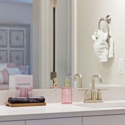 White bathroom vanity with a marble countertop, two gold faucets, and pink textured soap dispensers beside a black towel on a wooden tray; towel rings hold white towels, and a large mirror reflects a softly-lit bedroom.