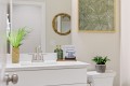 Modern bathroom vanity with white countertop, chrome faucet, and greenery in woven pot; wall art above and a small plant on the toilet tank; sign says ‘PLEASE USE THE MAKEUP TOWELS’.
