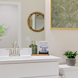 Modern bathroom vanity with white countertop, chrome faucet, and greenery in woven pot; wall art above and a small plant on the toilet tank; sign says ‘PLEASE USE THE MAKEUP TOWELS’.