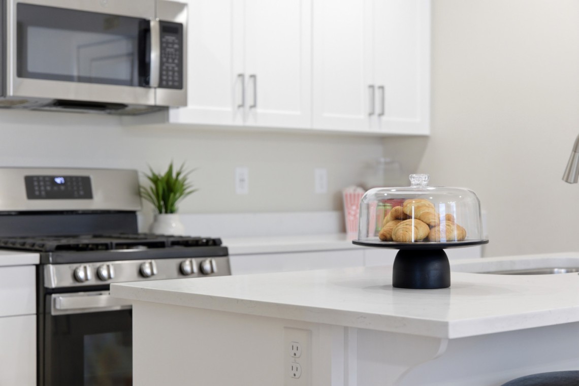 Cookies under a glass cloche on a black pedestal sits on a white kitchen island, with a stainless stove and microwave in the background.