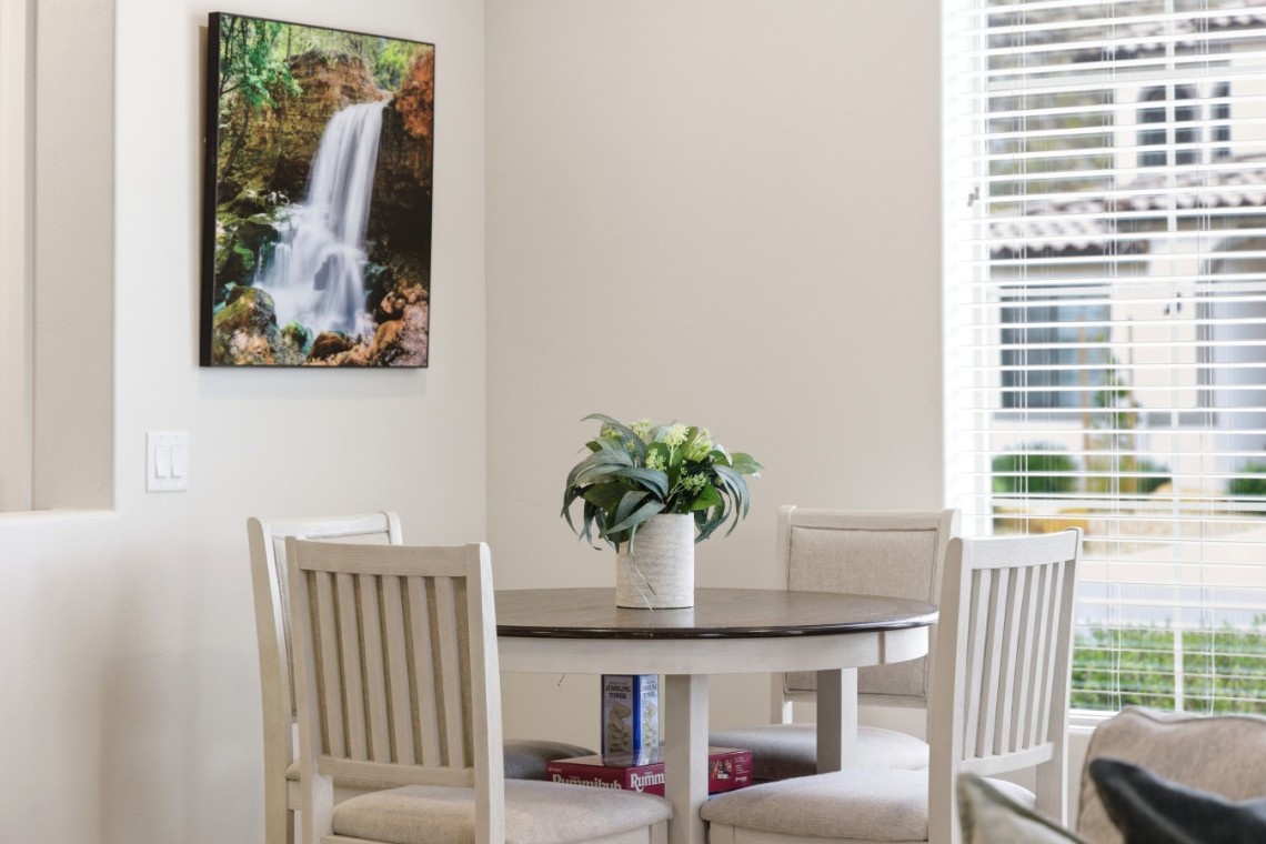 Round wooden dining table with a green plant centerpiece and four light chairs in a bright room; wall art of a waterfall on the left and large window blinds on the right.