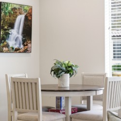 Round wooden dining table with a green plant centerpiece and four light chairs in a bright room; wall art of a waterfall on the left and large window blinds on the right.