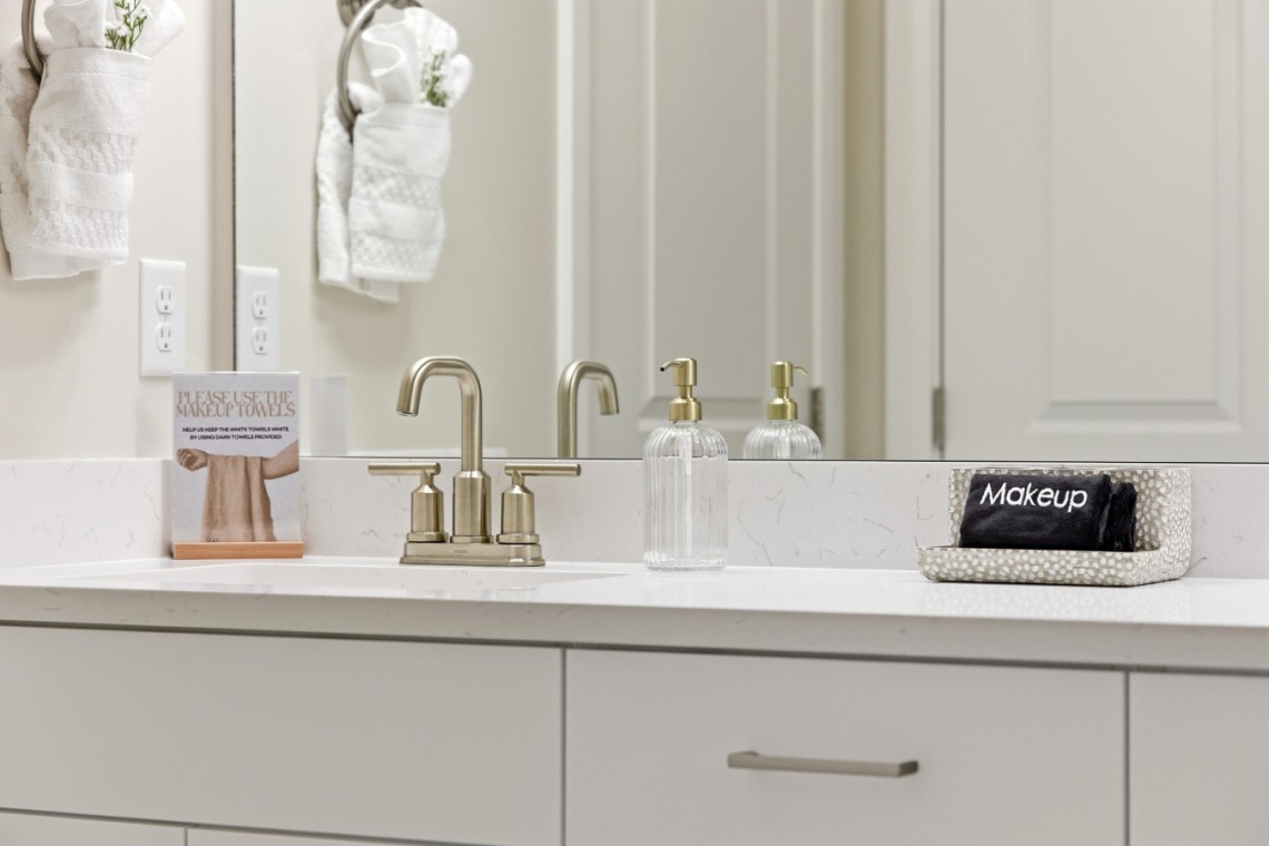 Hotel bathroom vanity with brushed-nickel faucet, clear soap dispenser, and a makeup-towel rack on the counter, plus a small card encouraging use of makeup towels and a black towel labeled 'Makeup' in a dotted tray.