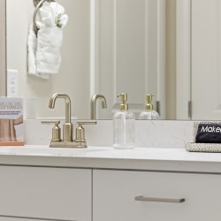 Hotel bathroom vanity with brushed-nickel faucet, clear soap dispenser, and a makeup-towel rack on the counter, plus a small card encouraging use of makeup towels and a black towel labeled 'Makeup' in a dotted tray.