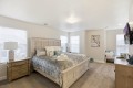 Bright neutral bedroom with a carved wooden bed, matching nightstands, blue-gray bedding, and a seating nook by windows.