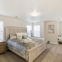 Bright neutral bedroom with a carved wooden bed, matching nightstands, blue-gray bedding, and a seating nook by windows.