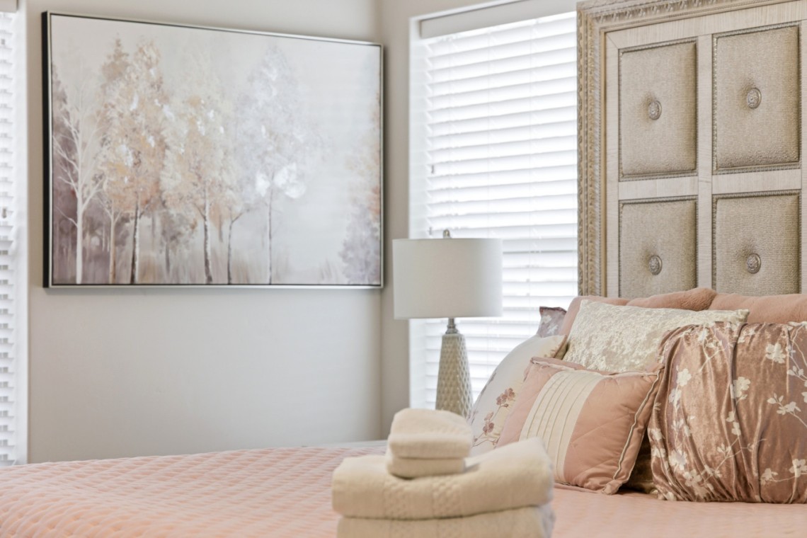 Blush pink bedroom with quilted bedspread, decorative pillows, stacked towels and a hat on the bed; beige tufted headboard and a framed tree artwork on the wall.
